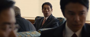 Movie still from “Shin Godzilla” (2016), directed by Shinji Higuchi – An older man in a suit and tie sitting in a chair; Close Up shot, Over the shoulder angle
