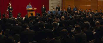 Movie still from “Shin Godzilla” (2016), directed by Shinji Higuchi – A group of men in suits are sitting in front of an audience; Extreme Wide shot, High angle