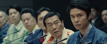Movie still from “Shin Godzilla” (2016), directed by Shinji Higuchi – A group of men sitting in front of microphones in a room; Close Up shot, Over the shoulder angle