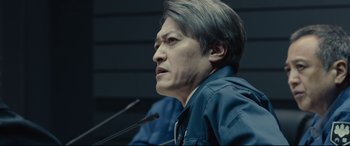 Movie still from “Shin Godzilla” (2016), directed by Shinji Higuchi – A man with short black hair is sitting in front of a microphone; Close Up shot, Low angle