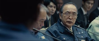 Movie still from “Shin Godzilla” (2016), directed by Shinji Higuchi – A group of men sitting next to each other in front of a microphone; Close Up shot, Over the shoulder angle