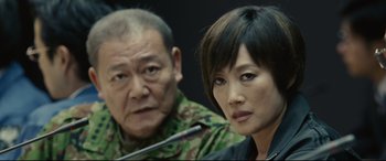 Movie still from “Shin Godzilla” (2016), directed by Shinji Higuchi – A man and a woman sitting next to each other in front of a microphone; Close Up shot, Low angle