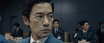 Movie still from “Shin Godzilla” (2016), directed by Shinji Higuchi – A man in a suit and glasses in front of another man in a suit and glasses; Close Up shot, Over the shoulder angle