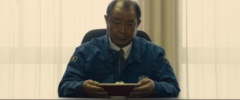 Movie still from “Shin Godzilla” (2016), directed by Shinji Higuchi – An older man sitting at a desk holding a book; Medium shot, Low angle