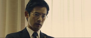 Movie still from “Shin Godzilla” (2016), directed by Shinji Higuchi – A person wearing glasses and a suit; Close Up shot, Over the shoulder angle