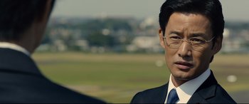 Movie still from “Shin Godzilla” (2016), directed by Shinji Higuchi – A person wearing a suit and tie; Close Up shot, Over the shoulder angle