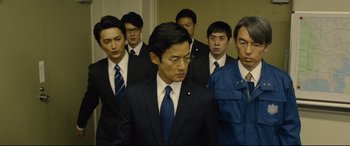 Movie still from “Shin Godzilla” (2016), directed by Shinji Higuchi – A group of men in suits and ties standing in a room; Medium shot, Low angle
