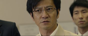 Movie still from “Shin Godzilla” (2016), directed by Shinji Higuchi – A person wearing glasses and a suit; Close Up shot, Low angle
