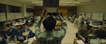 Movie still from “Shin Godzilla” (2016), directed by Shinji Higuchi – A man holding up an open laptop computer in front of a crowd; Wide shot, Overhead angle