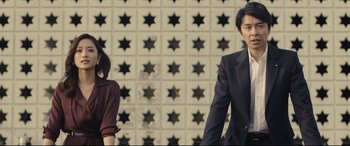 Movie still from “Shin Godzilla” (2016), directed by Shinji Higuchi – A man and a woman standing next to each other in front of a wall; Medium shot, Low angle