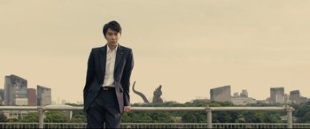 Movie still from “Shin Godzilla” (2016), directed by Shinji Higuchi – A man in a suit standing in front of a statue; Medium shot, Low angle