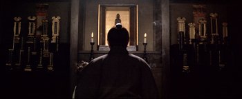 Movie still from “Shogun Assassin” (1980), directed by Kenji Misumi – A man standing in front of candles in front of a statue; Medium shot, Low angle