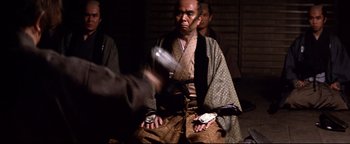 Movie still from “Shogun Assassin” (1980), directed by Kenji Misumi – A man in traditional japanese garb sitting on the ground; Medium shot, Over the shoulder angle