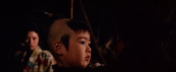 Movie still from “Shogun Assassin” (1980), directed by Kenji Misumi – A young boy with a shaved head sitting in a chair; Close Up shot, High angle