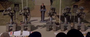 Movie still from “Short Circuit” (1986), directed by John Badham – A man is standing at a podium in front of a group of robots; Wide shot, High angle