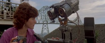 Movie still from “Short Circuit” (1986), directed by John Badham – A woman standing next to a camera on top of a building; Close Up shot, Low angle