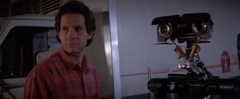 Movie still from “Short Circuit” (1986), directed by John Badham – A man standing in front of a camera in front of a mirror; Close Up shot, Low angle