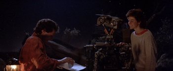 Movie still from “Short Circuit” (1986), directed by John Badham – Two people are looking at a robot on a table; Medium shot, Over the shoulder angle