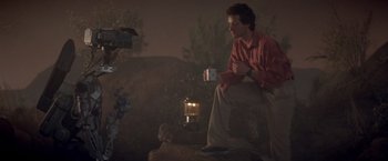 Movie still from “Short Circuit” (1986), directed by John Badham – A man sitting on the ground holding a cup; Wide shot, Low angle