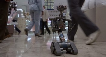 Movie still from “Short Circuit 2” (1988), directed by Kenneth Johnson – A robot is sitting on the ground in a mall; Wide shot, High angle