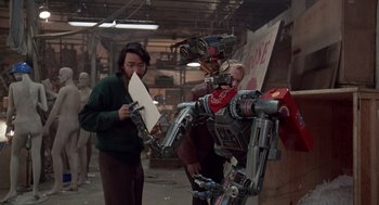 Movie still from “Short Circuit 2” (1988), directed by Kenneth Johnson – Two people looking at a robot in a room; Medium shot, Over the shoulder angle