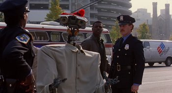 Movie still from “Short Circuit 2” (1988), directed by Kenneth Johnson – Two police officers standing next to a man with a robot head; Medium shot, Over the shoulder angle