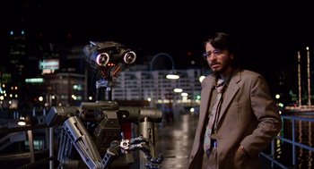 Movie still from “Short Circuit 2” (1988), directed by Kenneth Johnson – A man standing next to a robot in a city; Medium shot, Over the shoulder angle