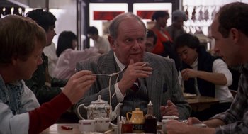 Movie still from “Short Circuit 2” (1988), directed by Kenneth Johnson – An older man sitting in front of a group of people; Medium shot, Over the shoulder angle