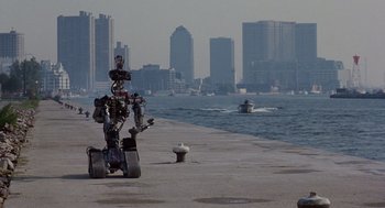Movie still from “Short Circuit 2” (1988), directed by Kenneth Johnson – A robot is sitting on a pier near the water; Extreme Wide shot, High angle