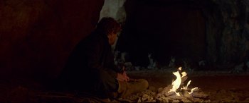 Movie still from “Siberia” (2020), directed by Abel Ferrara – A man sitting on the ground near a fire pit; Wide shot, Over the shoulder angle