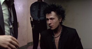 Movie still from “Sid and Nancy” (1986), directed by Alex Cox – A man in a black suit and a police officer in the background; Medium shot, Low angle