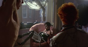 Movie still from “Sid and Nancy” (1986), directed by Alex Cox – A woman sitting in a chair reading a magazine; Wide shot, Over the shoulder angle