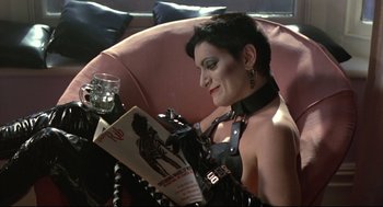 Movie still from “Sid and Nancy” (1986), directed by Alex Cox – A woman sitting in a chair reading a magazine; Medium shot, High angle