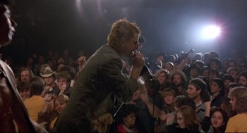 Movie still from “Sid and Nancy” (1986), directed by Alex Cox – A man is singing into a microphone in front of a crowd; Wide shot, Low angle