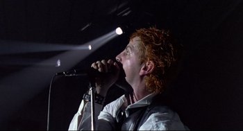 Movie still from “Sid and Nancy” (1986), directed by Alex Cox – A man with curly red hair singing into a microphone; Close Up shot, Low angle
