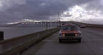 Movie still from “Sid and Nancy” (1986), directed by Alex Cox – A car driving down a road next to a body of water; Extreme Wide shot, Low angle