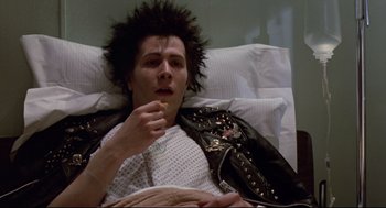 Movie still from “Sid and Nancy” (1986), directed by Alex Cox – A man in a white shirt and a black jacket is laying in a hospital bed; Medium shot, Low angle
