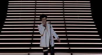Movie still from “Sid and Nancy” (1986), directed by Alex Cox – A man standing on a stage holding a microphone; Wide shot, Overhead angle