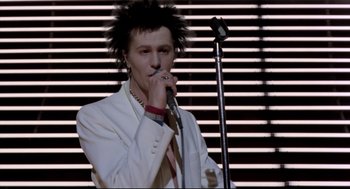Movie still from “Sid and Nancy” (1986), directed by Alex Cox – A man in a white suit is singing into a microphone; Medium shot, Low angle