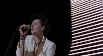 Movie still from “Sid and Nancy” (1986), directed by Alex Cox – A man in a white jacket is singing into a microphone; Medium shot, Low angle