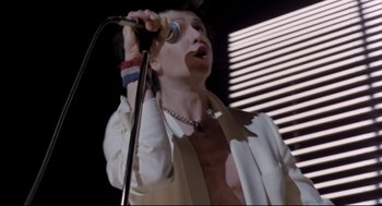 Movie still from “Sid and Nancy” (1986), directed by Alex Cox – A man in a white shirt is singing into a microphone; Medium shot, Low angle