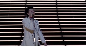 Movie still from “Sid and Nancy” (1986), directed by Alex Cox – A man in a white jacket is singing into a microphone; Wide shot, Low angle
