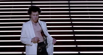Movie still from “Sid and Nancy” (1986), directed by Alex Cox – A man in a white jacket and black pants standing in front of a window; Medium shot, Low angle