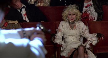 Movie still from “Sid and Nancy” (1986), directed by Alex Cox – A woman sitting on a red chair wearing a white dress; Medium shot, Over the shoulder angle