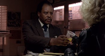 Movie still from “Sid and Nancy” (1986), directed by Alex Cox – A man sitting at a table holding a cell phone; Close Up shot, Low angle