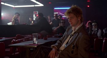 Movie still from “Sid and Nancy” (1986), directed by Alex Cox – A man sitting at a table in front of a group of people; Medium shot, Over the shoulder angle