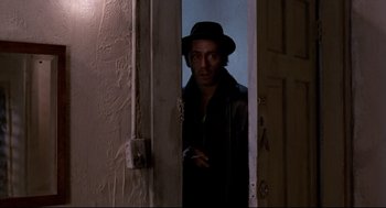 Movie still from “Sid and Nancy” (1986), directed by Alex Cox – A man wearing a hat standing in front of an open door; Medium shot, Over the shoulder angle