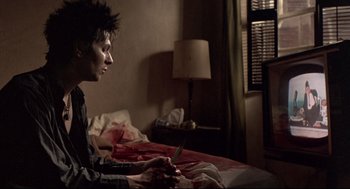 Movie still from “Sid and Nancy” (1986), directed by Alex Cox – A man sitting on a bed holding a knife; Medium shot, Over the shoulder angle