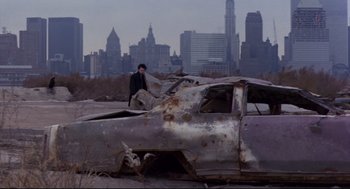 Movie still from “Sid and Nancy” (1986), directed by Alex Cox – A man standing next to an old car in a field; Wide shot, High angle