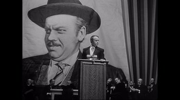 Movie still from “Side by Side” (2012), directed by Christopher Kenneally – A man in a suit and tie standing at a podium in front of a large picture of a man; Medium shot, Low angle